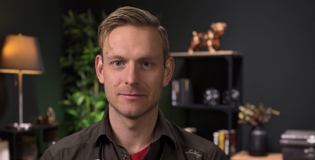 En instruktør i mørk skjorte ser rett inn i kameraet i et varmt innemiljø.